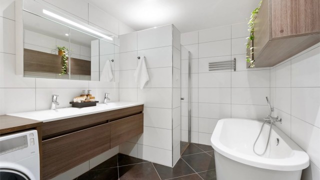 Badet har vegghengt toalett, vegghengt servantskap med dobbel nedsenket servant, vegghengt speilskap med overbelysning og dusjsone med innfellbare glassdører, hånddusj og regndusj, samt badekar