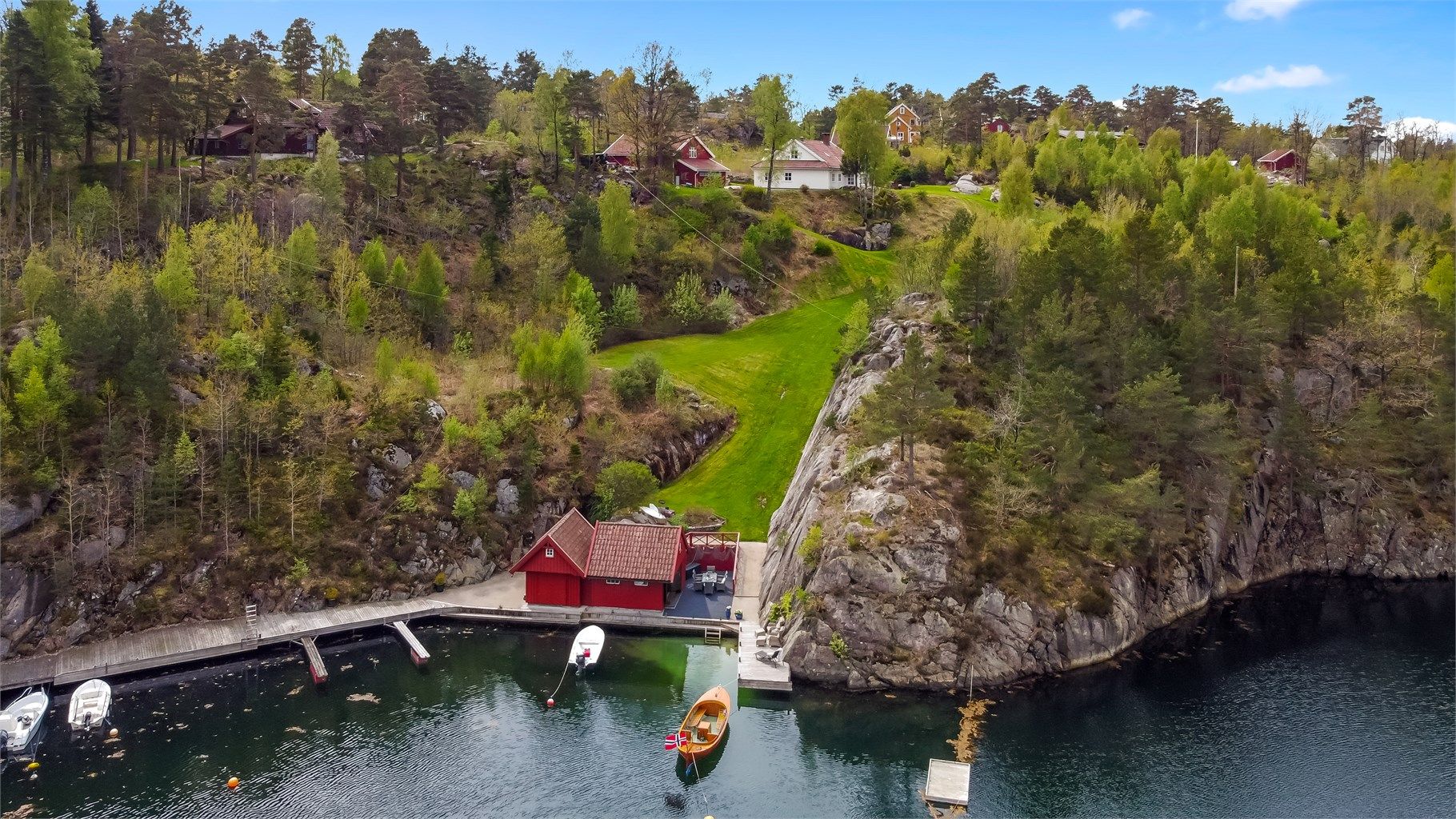 Oversiktsbilde fra sjøboden med brygge og tilhørende gressplen som fører opp til bolig og garasje 
