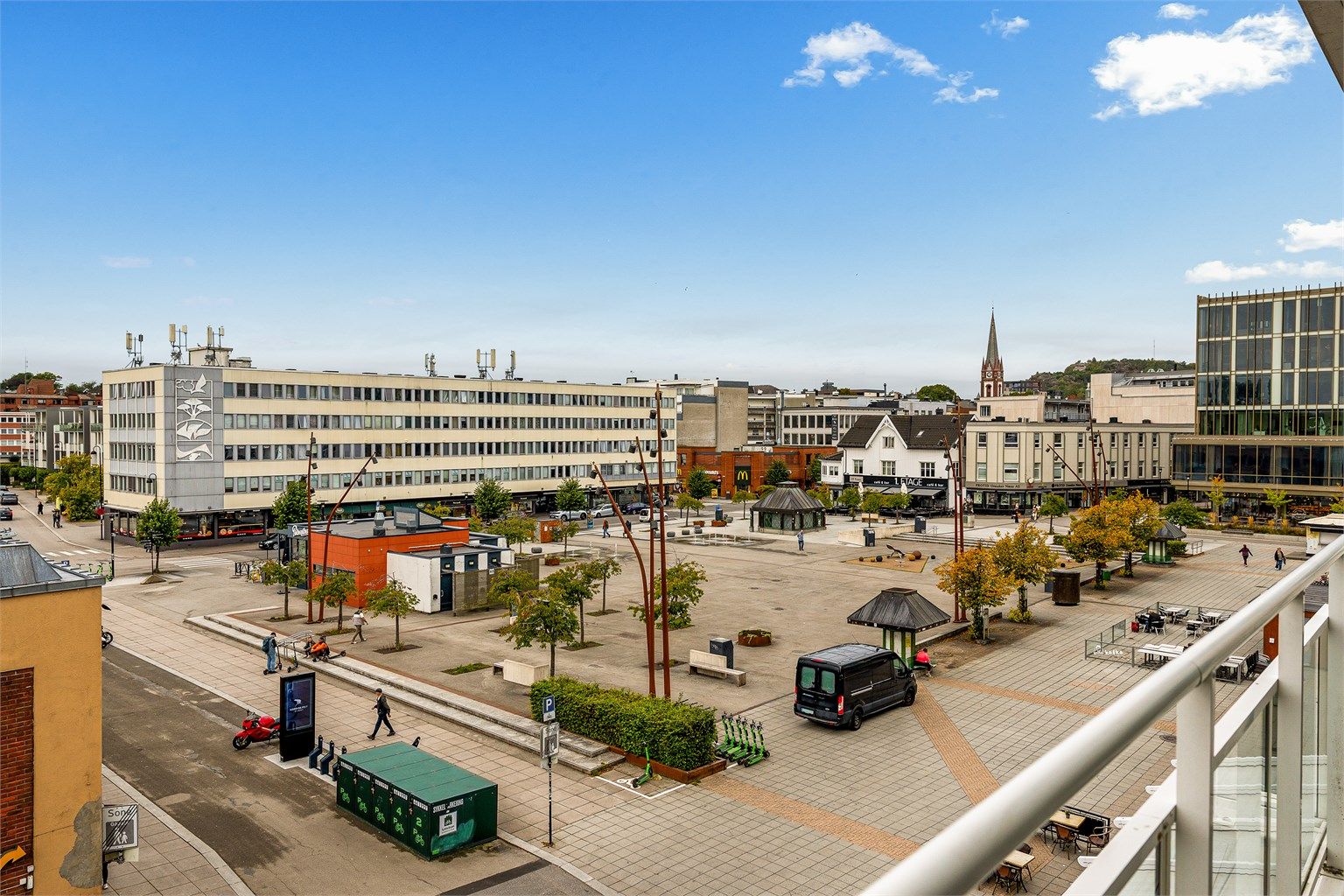 Åpent utsyn over Torget med byliv og kirketårnet i bakgrunnen.