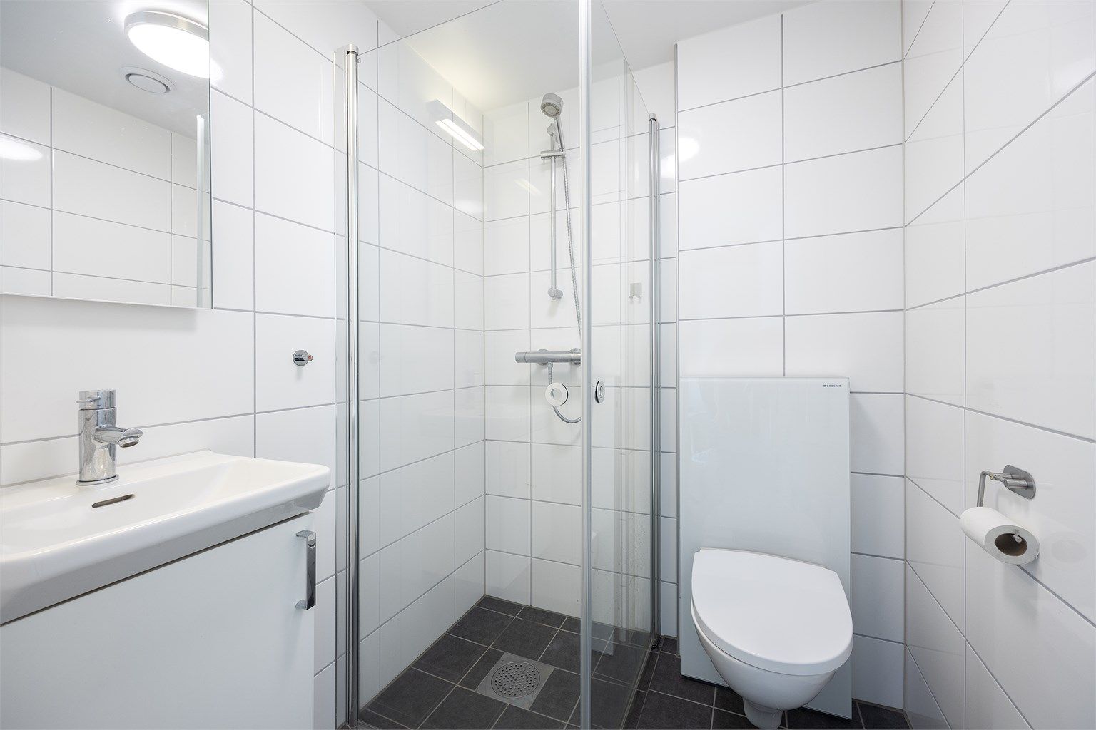 Badet fra soverom III / hybeldelen er lyst og hyggelig med veggmontert toalett og speil, heldekkende servant med underskap og dusjhjørne med dører i herdet glass