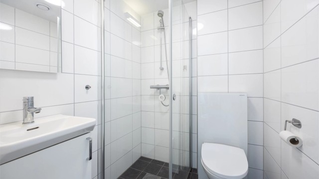 Badet fra soverom III / hybeldelen er lyst og hyggelig med veggmontert toalett og speil, heldekkende servant med underskap og dusjhjørne med dører i herdet glass