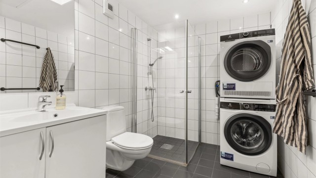 Flislagt baderom med elektrisk gulvvarme, dusjhjørne med glassdører og opplegg for vaskemaskin – praktisk utformet for en enkel hverdag.