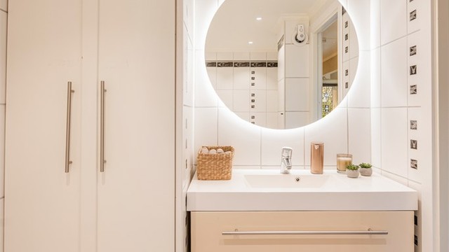 Vegghengt servantinnredning med romslige skuffer og rundt speil med integrert lys gir badet et stilrent og moderne preg, med høyskap for ekstra oppbevaring. Varmtvannstank i høyskap.
