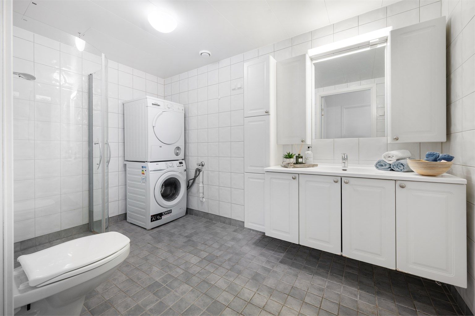 Badet har flislagt gulv med gulvvarme. Flislagte vegger. Takplater i himling. Vegghengt servantinnredning.