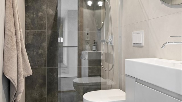 Badet har flislagte gulv og vegger, innredet med servant, vegghengt toalett og dusjhjørne.