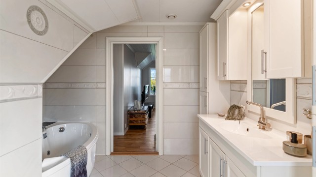 Det er montert vegghengte skap og baderomsinnredning med heldekkende servant. Speil, skap, overlys og stikkontakt er montert på vegg over servanten. Badekar og vegghengt toalett.