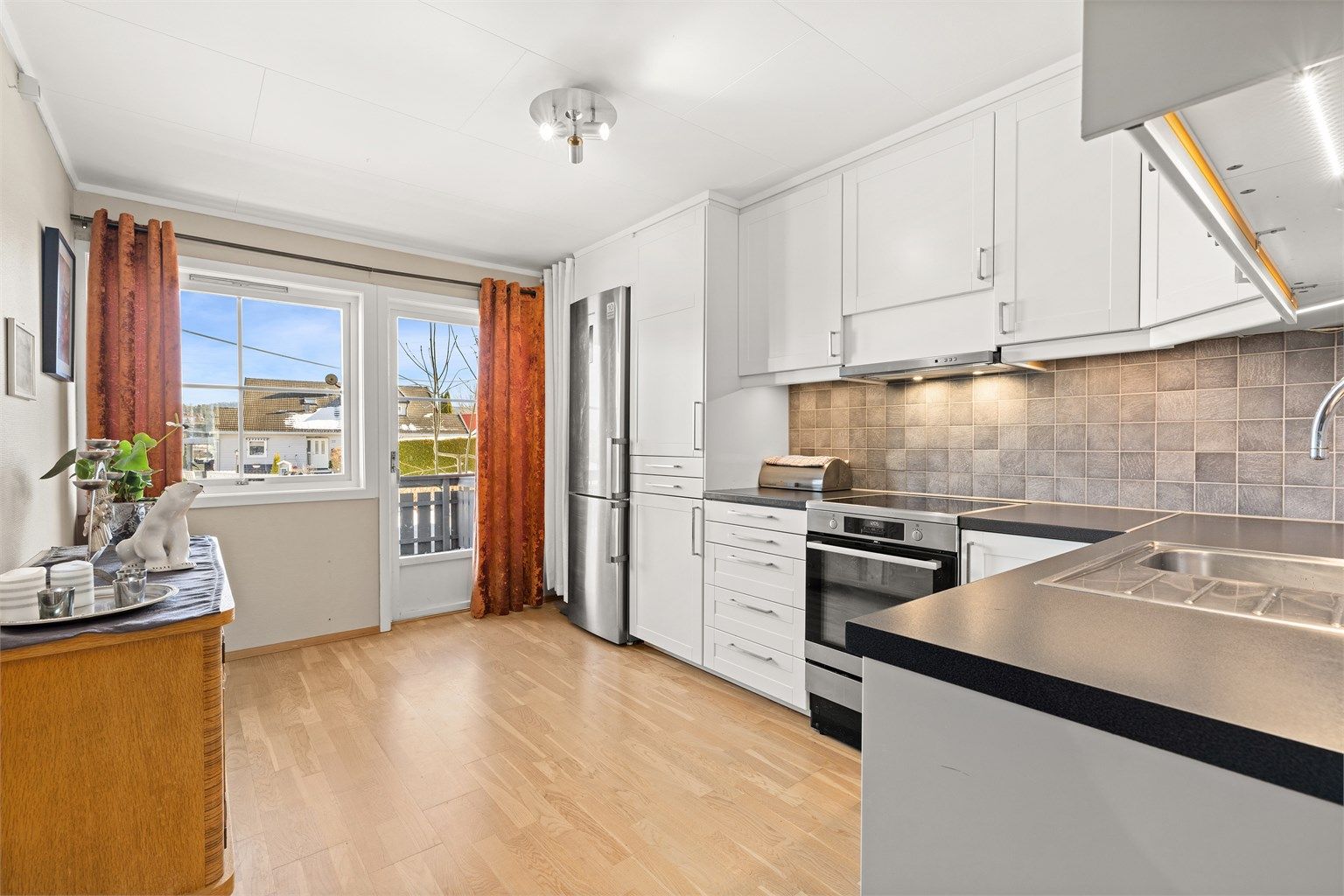 Romslig kjøkken med god benkeplass. Fliser på vegg mellom kjøkkenbenk og overskap. 