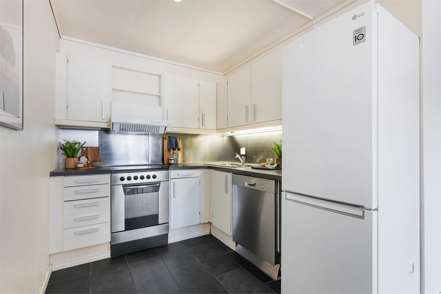 Kjøkkeninnredning med profilerte fronter og benkeplate med laminert overflate. Frittstående komfyr, oppvaskmaskin og kjøleskap med fryser. Ventilator i overskap fungerer ikke.