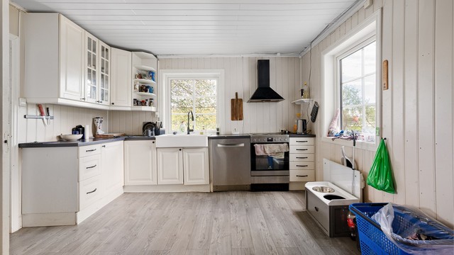 Innredning med profilerte fronter og laminert benkeplate med dobbel keramisk kum. Overskap med belysning, fritthengende ventilator med kullfilter og 3 doble stikkontakter. Hvitevarer som komfyr i nisje, innebygget oppvaskmaskin og kjøl/frys i nisje