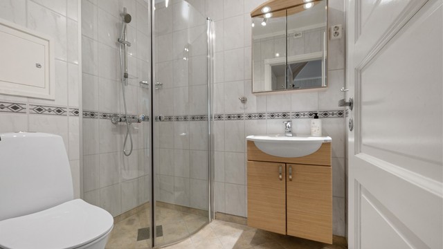 Badet har flislagt gulv med elektrisk gulvvarme. Den praktiske innredningen består av et vegghengt servantskap med heldekkende servant og ett-greps armatur, samt overskap med speildører.