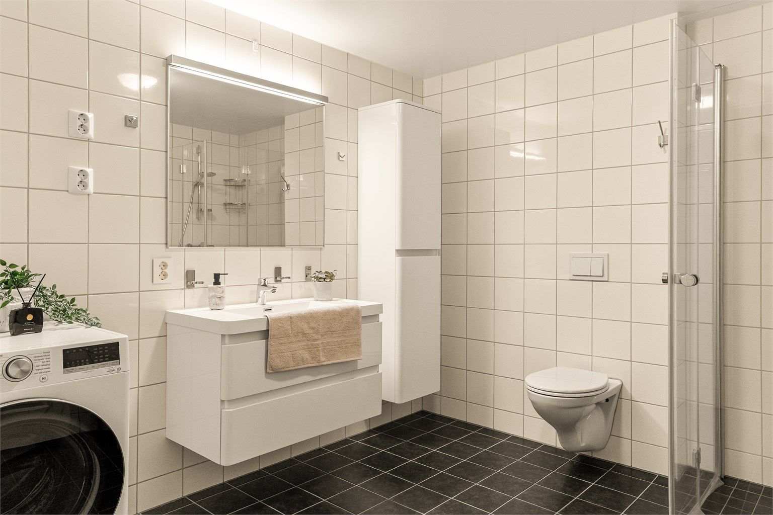 Pent flislagt bad med vegghengt toalett, foldbare dusjdører og god oppbevaring. Opplegg for vaskemaskin og plass til tørketrommel i rommet.