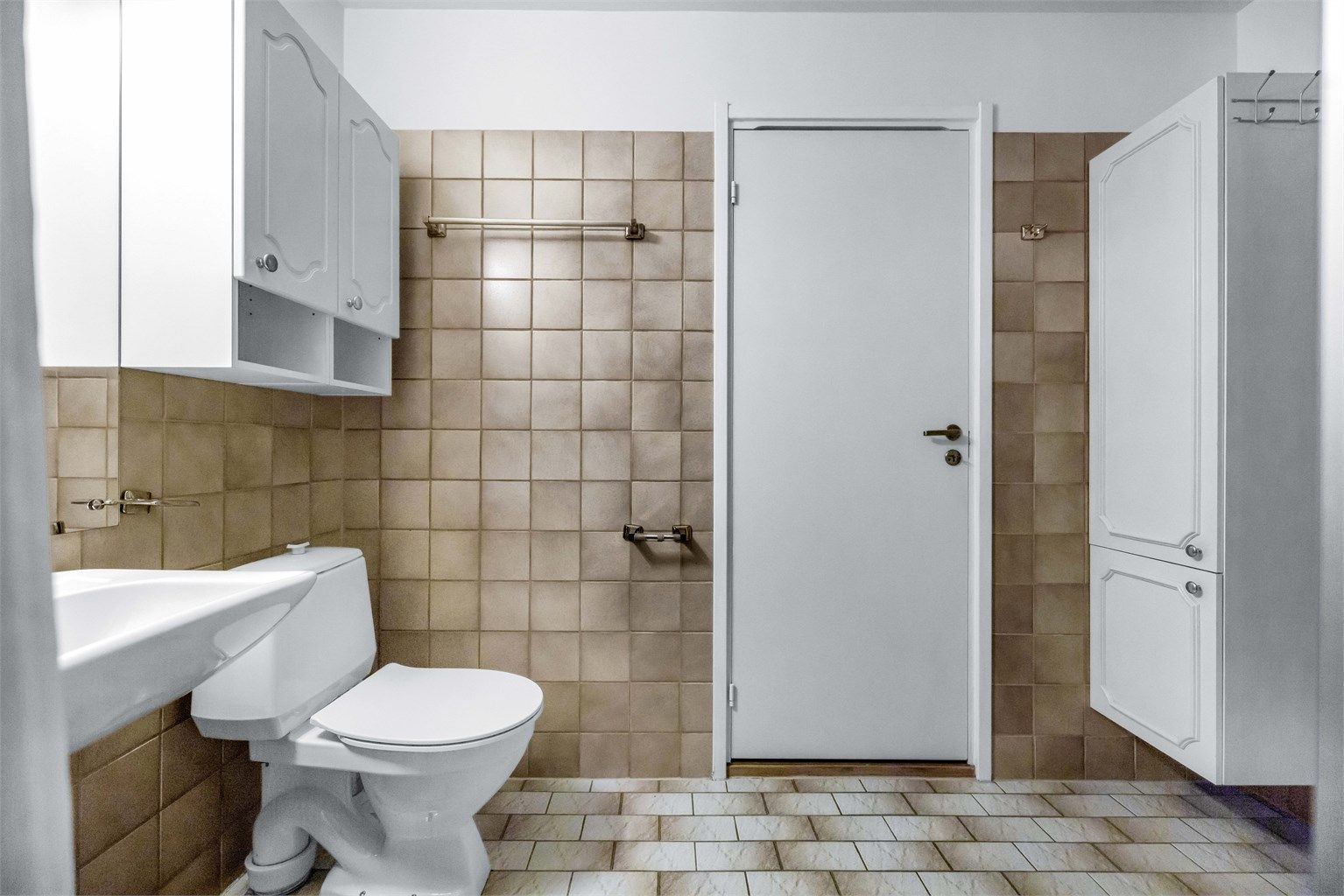 Badet er innredet med dusj, gulvstående toalett, vegghengt servant med armatur., speil og baderomsinnredning med hvite profilerte fronter. 