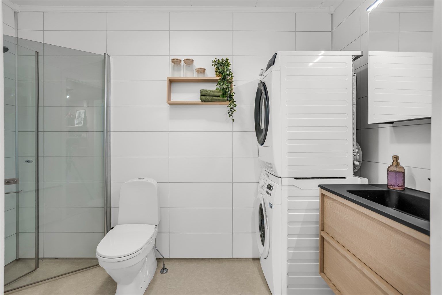 Badet har dusjhjørne med glassdører, hånddusj og frittstående toalett. Gulv med gulvbelegg og våtromsplater på veggene.