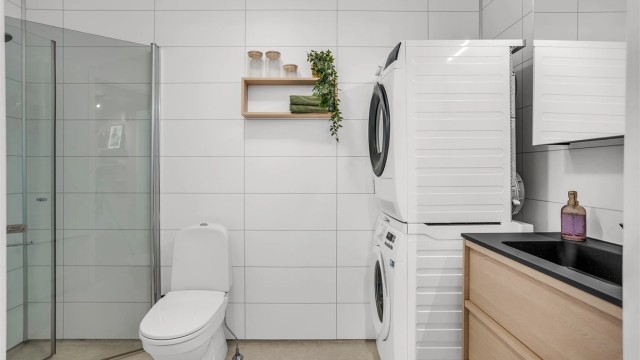 Badet har dusjhjørne med glassdører, hånddusj og frittstående toalett. Gulv med gulvbelegg og våtromsplater på veggene.