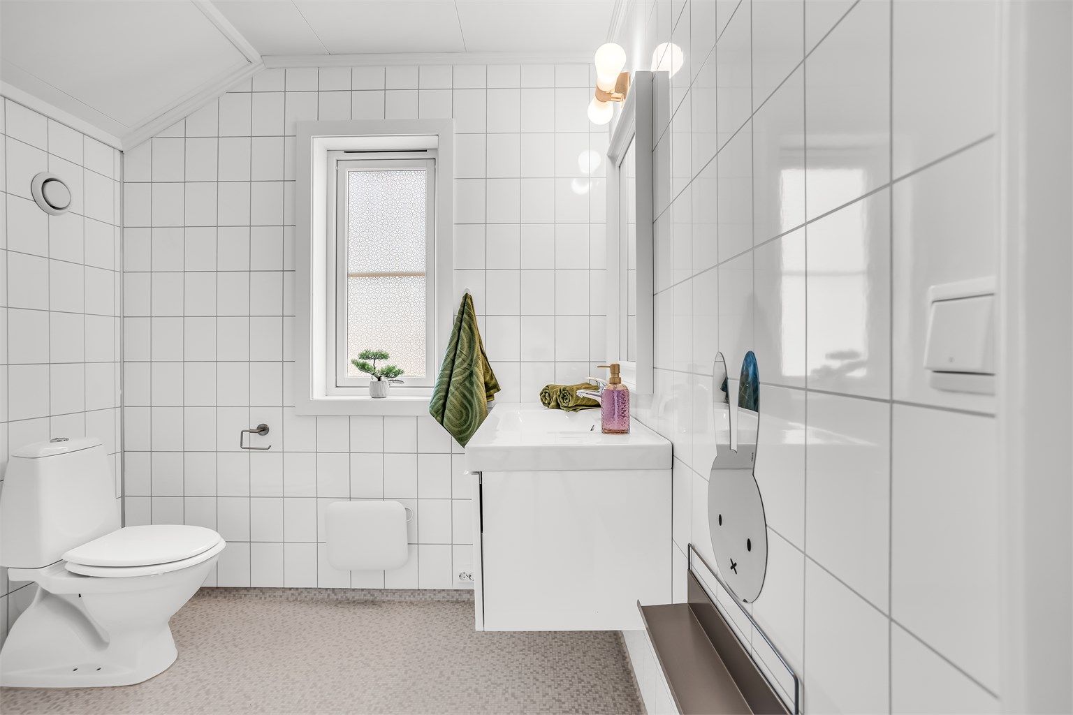 Bad i loftsetasjen med servantinnredning, speil med belysning og frittstående toalett. Gulv med gulvbelegg og våtromsplater på veggene.