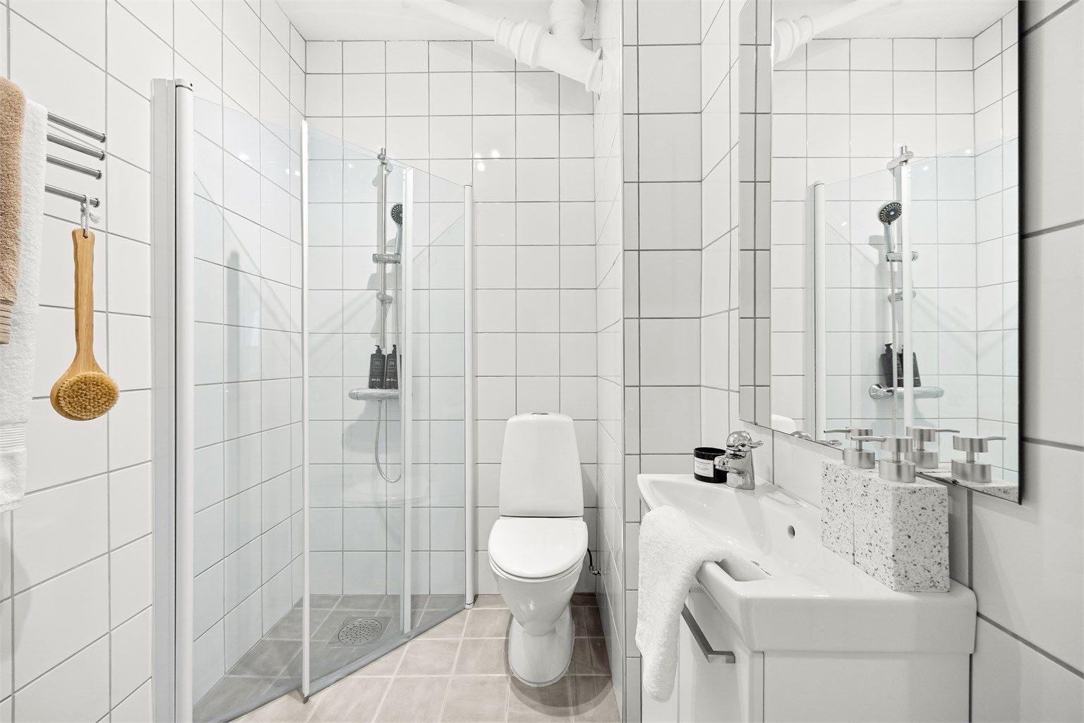 Badet er utstyrt med gulvstående toalett, vegghengt servantinnredning med ovenpåliggende vask og dusjnisje med dører.