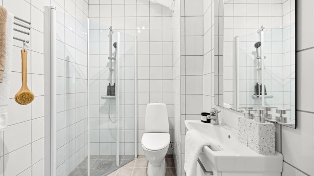 Badet er utstyrt med gulvstående toalett, vegghengt servantinnredning med ovenpåliggende vask og dusjnisje med dører.