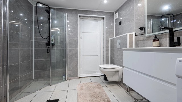 Badet har dusjhjørne, vegghengt toalett og baderomsplater på veggene.
