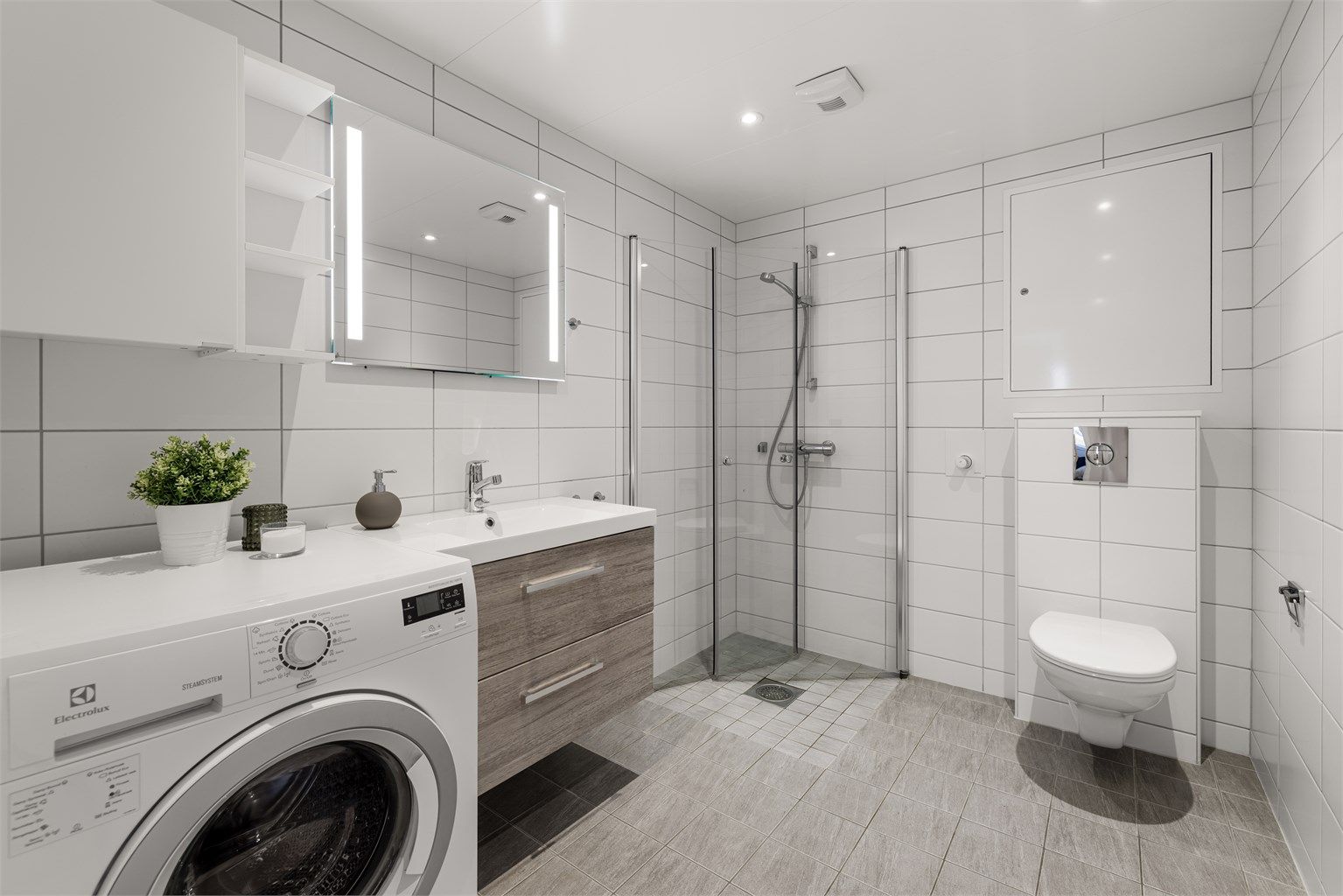 Badet er innredet med vegghengt servantinnredning, speil med sidelys, gulvstående høyskap og vegghengt toalett. Videre er det dusjhjørne med glassdører og vegghengt dusjarmatur. Det er også praktisk opplegg for vaskemaskin.