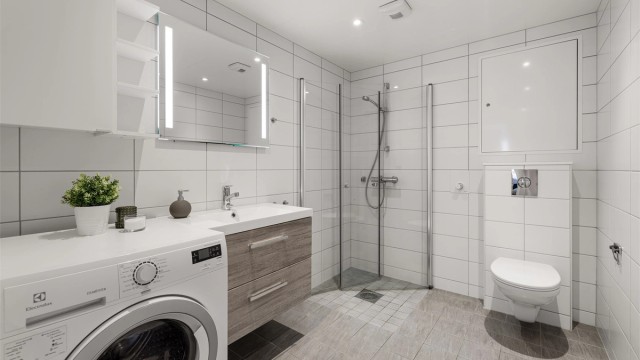 Badet er innredet med vegghengt servantinnredning, speil med sidelys, gulvstående høyskap og vegghengt toalett. Videre er det dusjhjørne med glassdører og vegghengt dusjarmatur. Det er også praktisk opplegg for vaskemaskin.