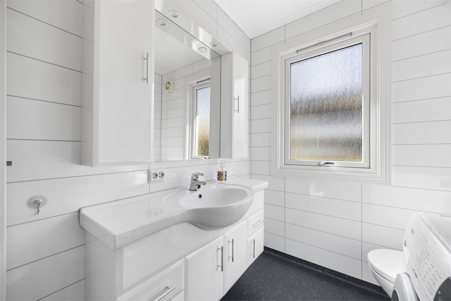 Baderomsinnredning med heldekkende servant, speil med overlys og et praktisk vegghengt skap.