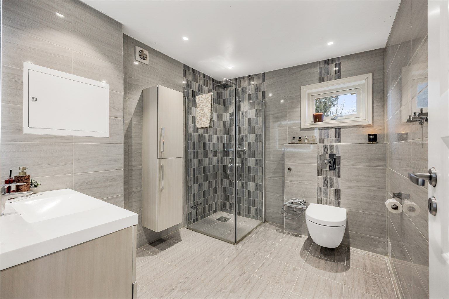 Rommet har vegghengt servantinnredning, speil med integrert lys og et dusjhjørne med innfellbare dører.