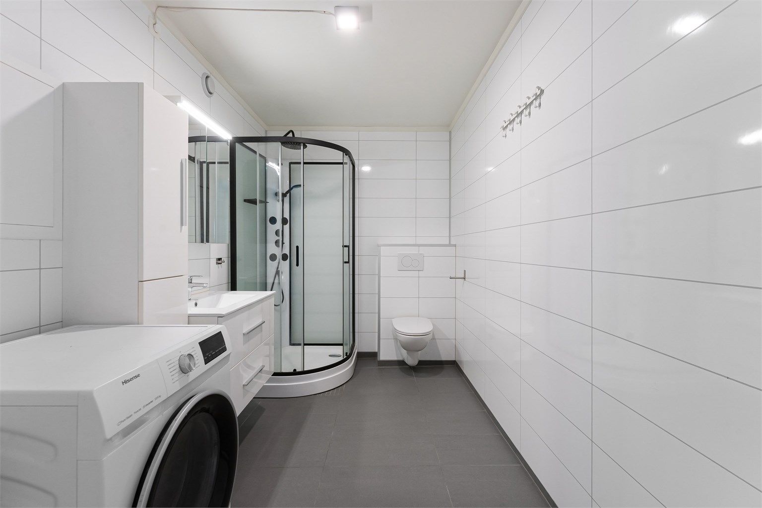 Badet er holdt i nøytrale farger med store, hvite veggfliser og mørkere gulvfliser.