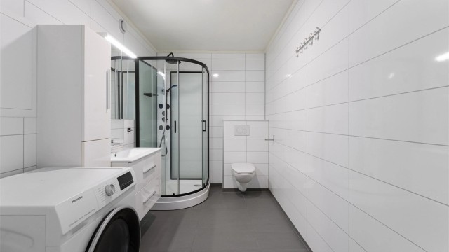 Badet er holdt i nøytrale farger med store, hvite veggfliser og mørkere gulvfliser.