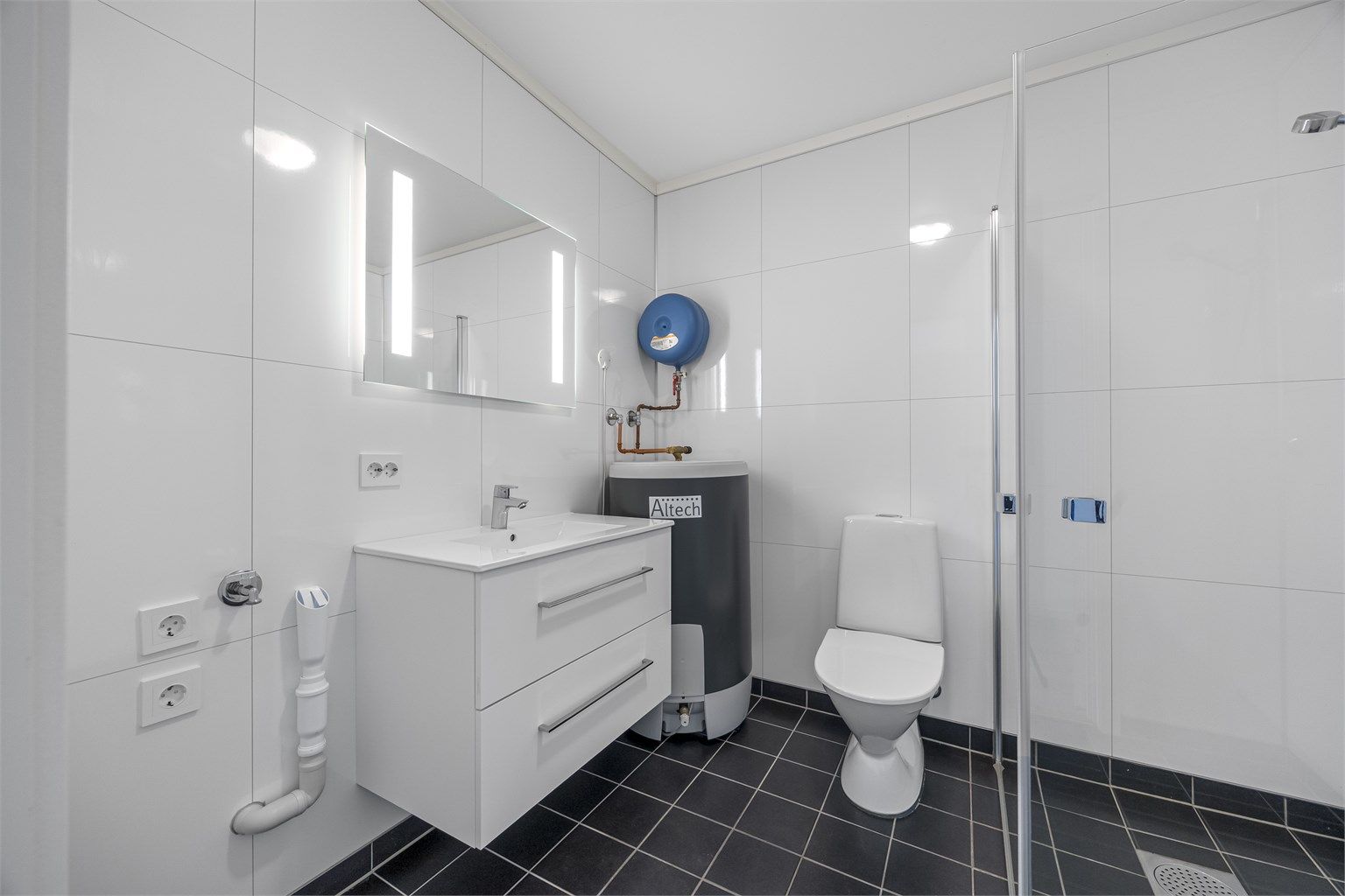 Baderom med sammenskyvbare dusjdører, opplegg til vaskemaskin, toalett, varmtvannstank, servant og speil med lys.