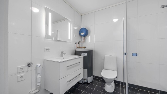 Baderom med sammenskyvbare dusjdører, opplegg til vaskemaskin, toalett, varmtvannstank, servant og speil med lys.