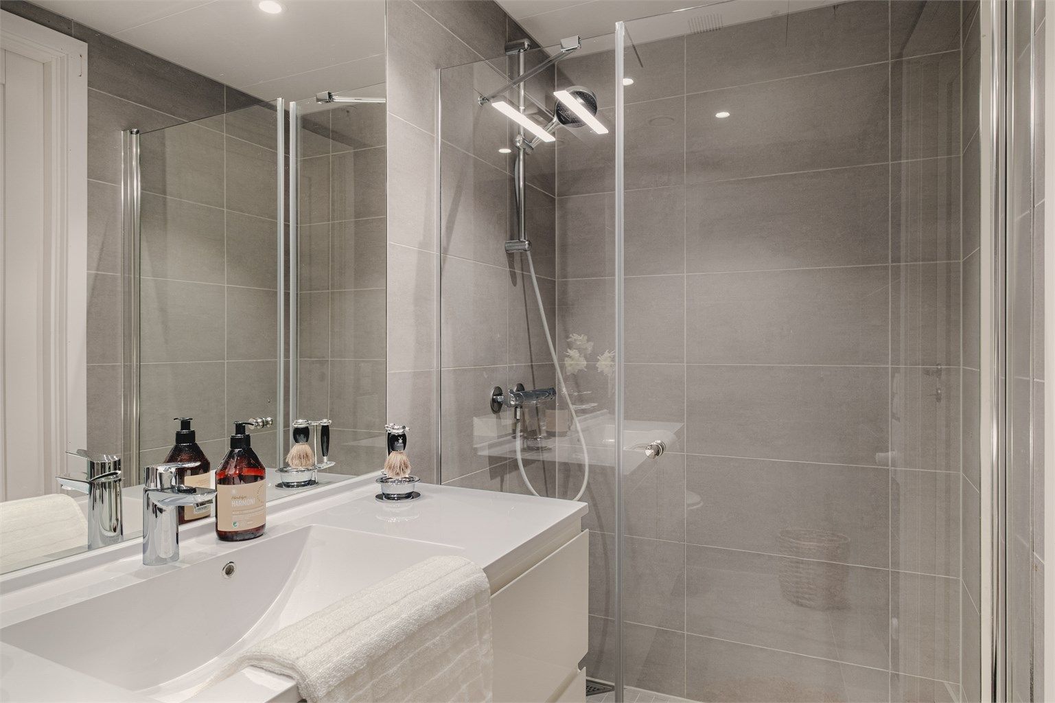 Badet er innredet med baderomsmøbler, speil i flukt med fliser og LED lyslist over, stor dusjnisje og vegghengt wc.