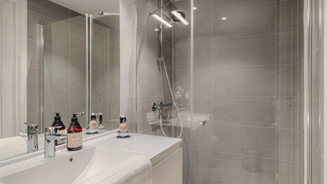 Badet er innredet med baderomsmøbler, speil i flukt med fliser og LED lyslist over, stor dusjnisje og vegghengt wc.