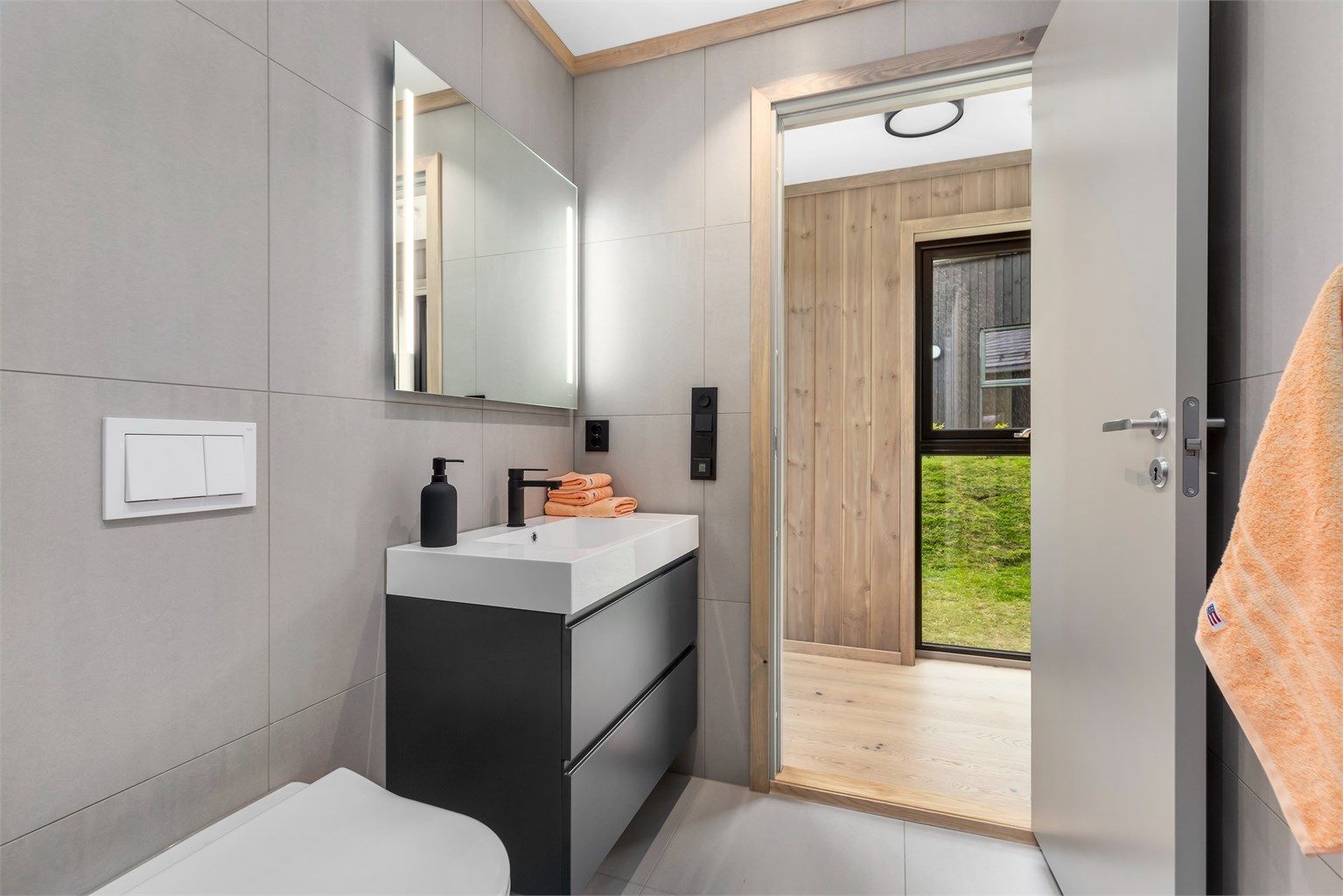 Badet er praktisk og delikat innredet med flislagt gulv og vegger. Heldekkende servant i seksjon med skuffer, speil med integrert belysning, wc med innebygget cisterne, dusjhjørne og opplegg til vaskemaskin plassert i en nisje.