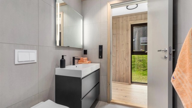 Badet er praktisk og delikat innredet med flislagt gulv og vegger. Heldekkende servant i seksjon med skuffer, speil med integrert belysning, wc med innebygget cisterne, dusjhjørne og opplegg til vaskemaskin plassert i en nisje.