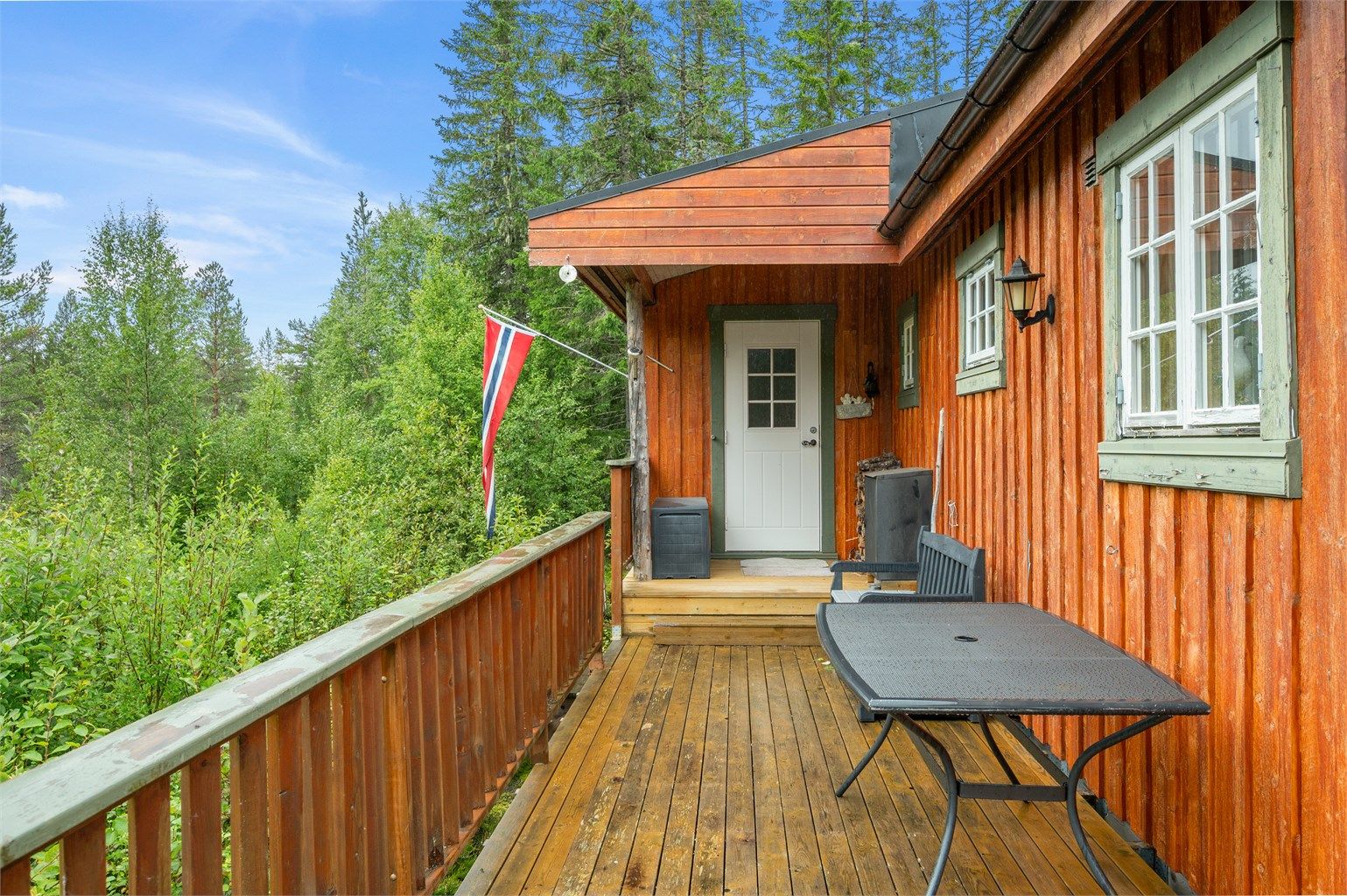 Hytta har et lunt og tradisjonelt uttrykk, med terrasse langs fasaden og grønne omgivelser på alle kanter.