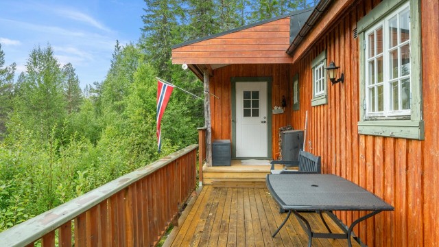 Hytta har et lunt og tradisjonelt uttrykk, med terrasse langs fasaden og grønne omgivelser på alle kanter.