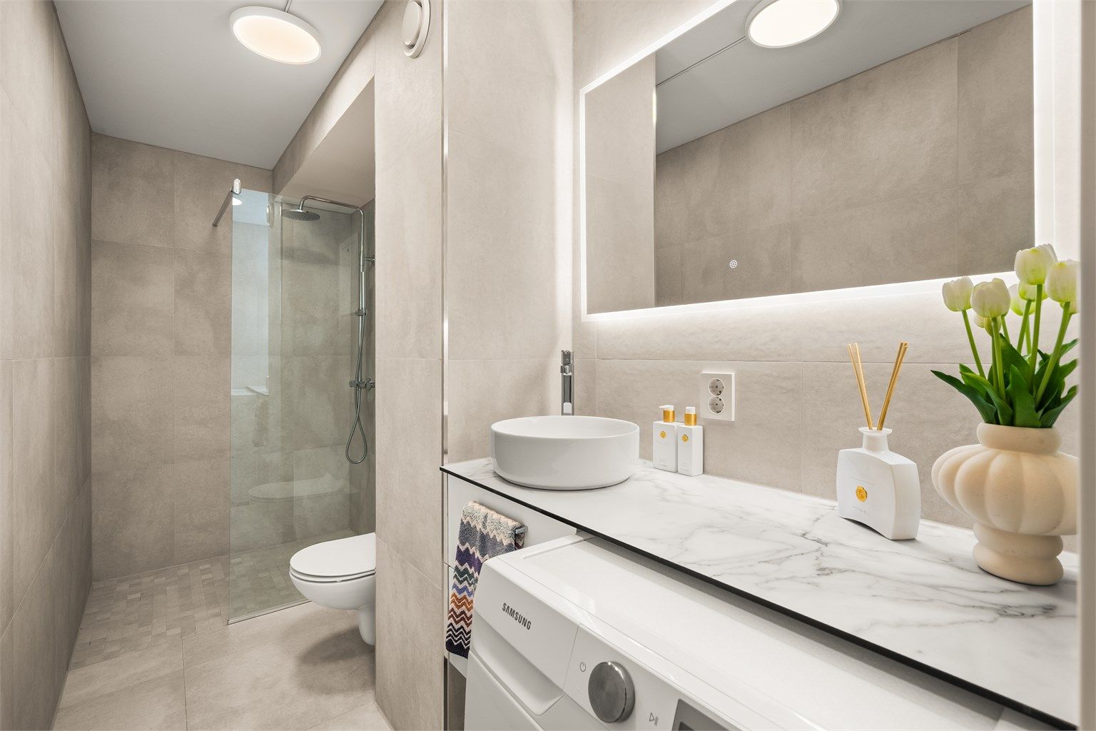 Lekkert, flislagt bad med gulvvarme fra 2025. Badet er innredet med vegghengt toalett, dusjhjørne med glassvegg og opplegg for vaskemaskin.