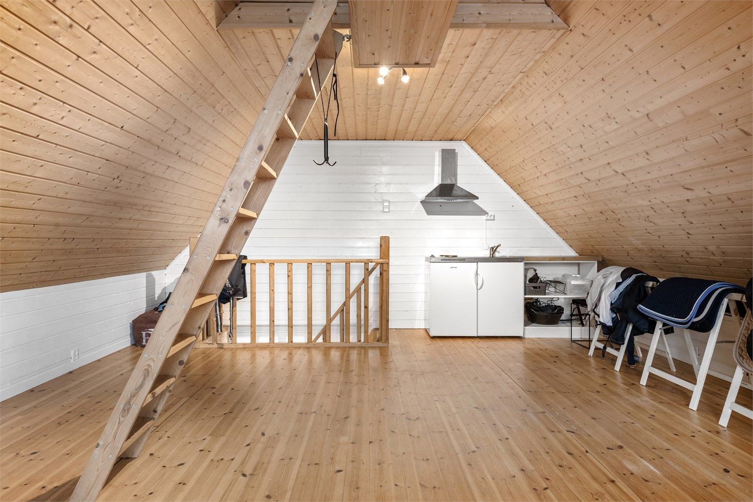 Innredet rom over garasjen med trebord på gulv, malt panel og et enkelt hybelkjøkken.