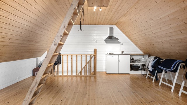 Innredet rom over garasjen med trebord på gulv, malt panel og et enkelt hybelkjøkken.