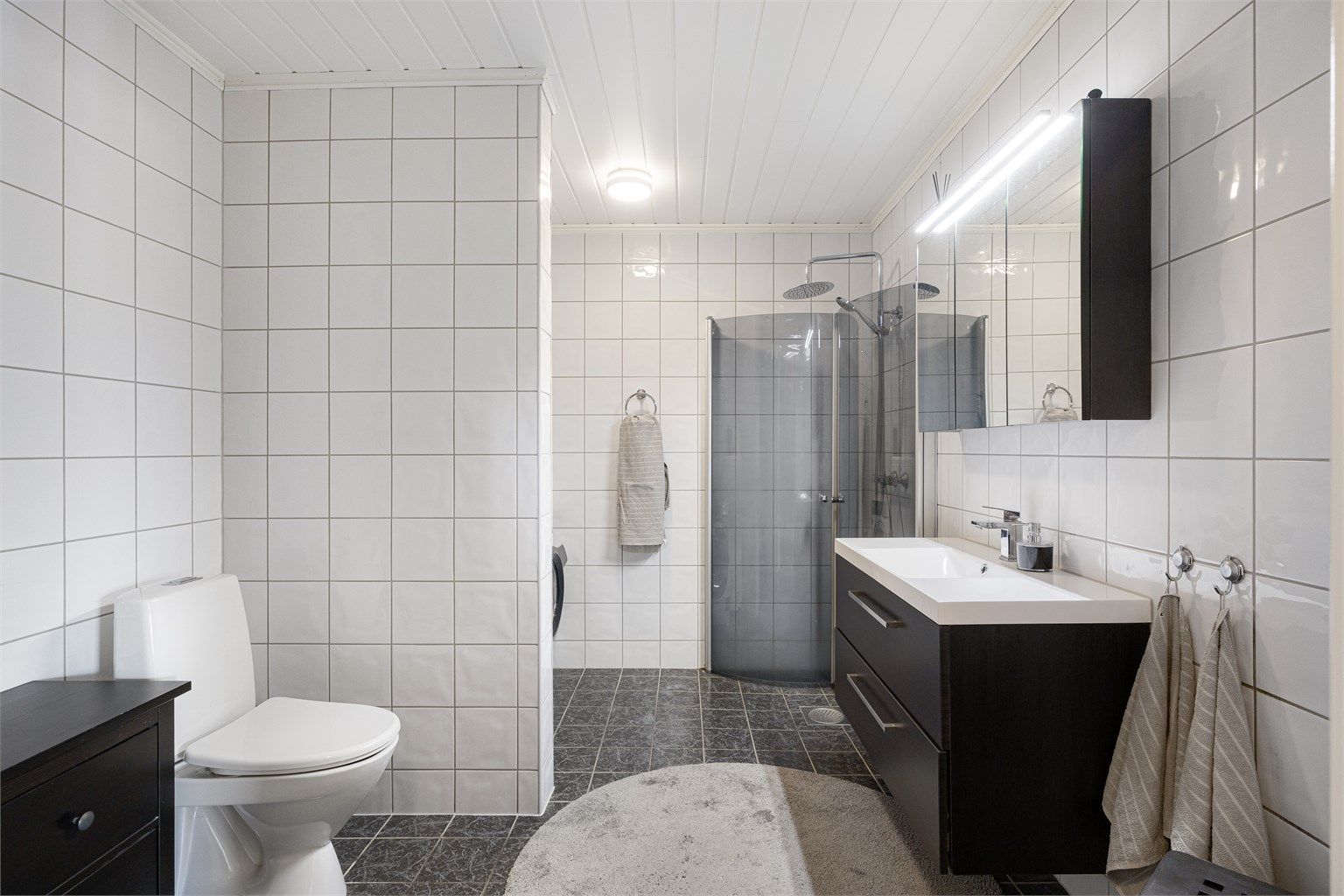 Romslig bad med flislagt gulv med varmekabler. Innredningen består av servantskap, speilskap og gulvmontert toalett.