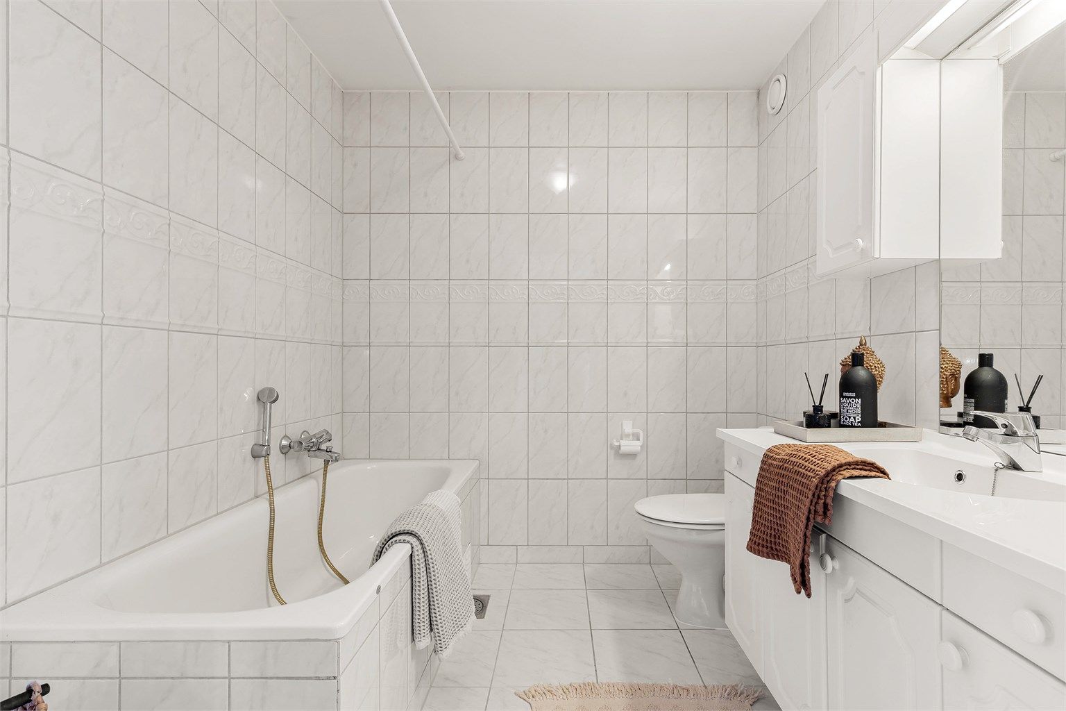 Badet har vegghengt servantinnredning med speil og overlys. Badekar med vegghengt badekararmatur og gulvstående toalett.