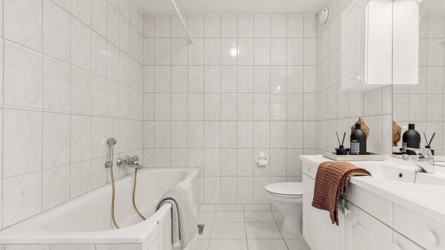 Badet har vegghengt servantinnredning med speil og overlys. Badekar med vegghengt badekararmatur og gulvstående toalett.