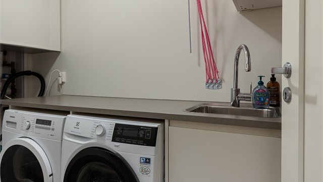 Praktisk vaskerom med opplegg for vaskemaskin og tørketrommel, utslagsvask, skapplass og tørkestativ i tak. Rommet har sluk i gulv.