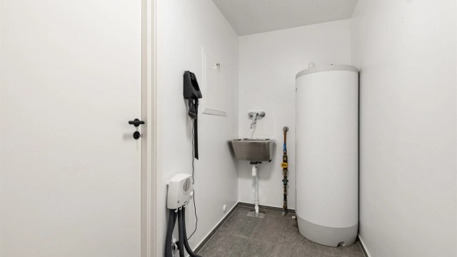 Praktisk vaskerom fra 2024 med flislagt gulv og gulvvarme. Vegghengt utslagsvask med ett-greps armatur, varmtvannsbereder og opplegg for vaskemaskin og tørketrommel.