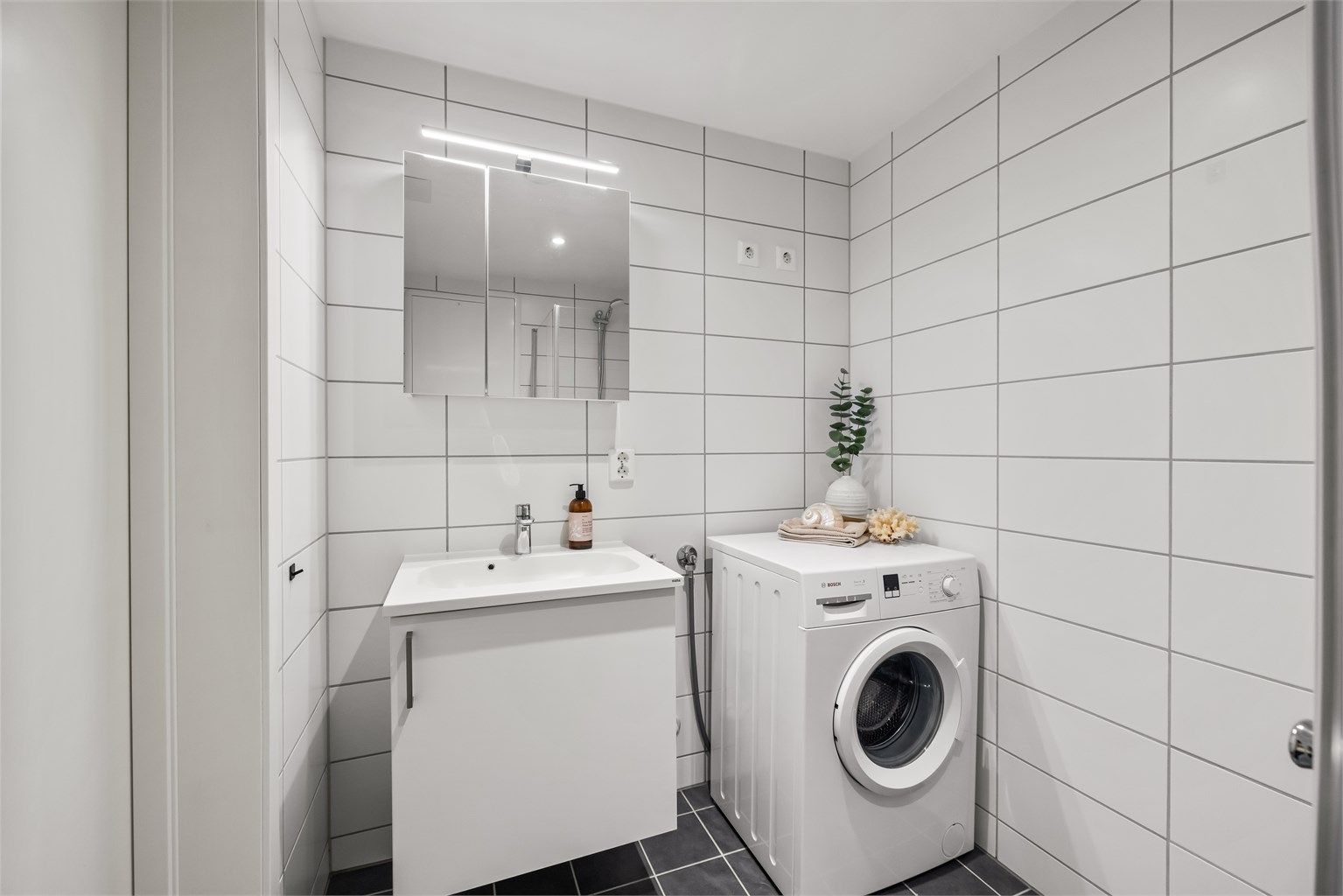 Badet er utstyrt med vegghengt toalett, dusjhjørne og servant med speilskap.