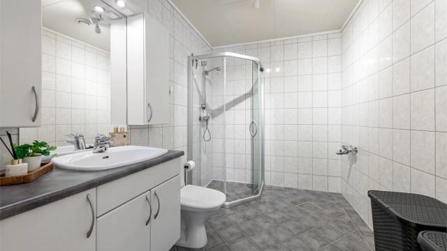 Badet har flislagt gulv med gulvvarme, flislagte vegger og et dusjhjørne med skyvedører. Rommet er klargjort for badekar.