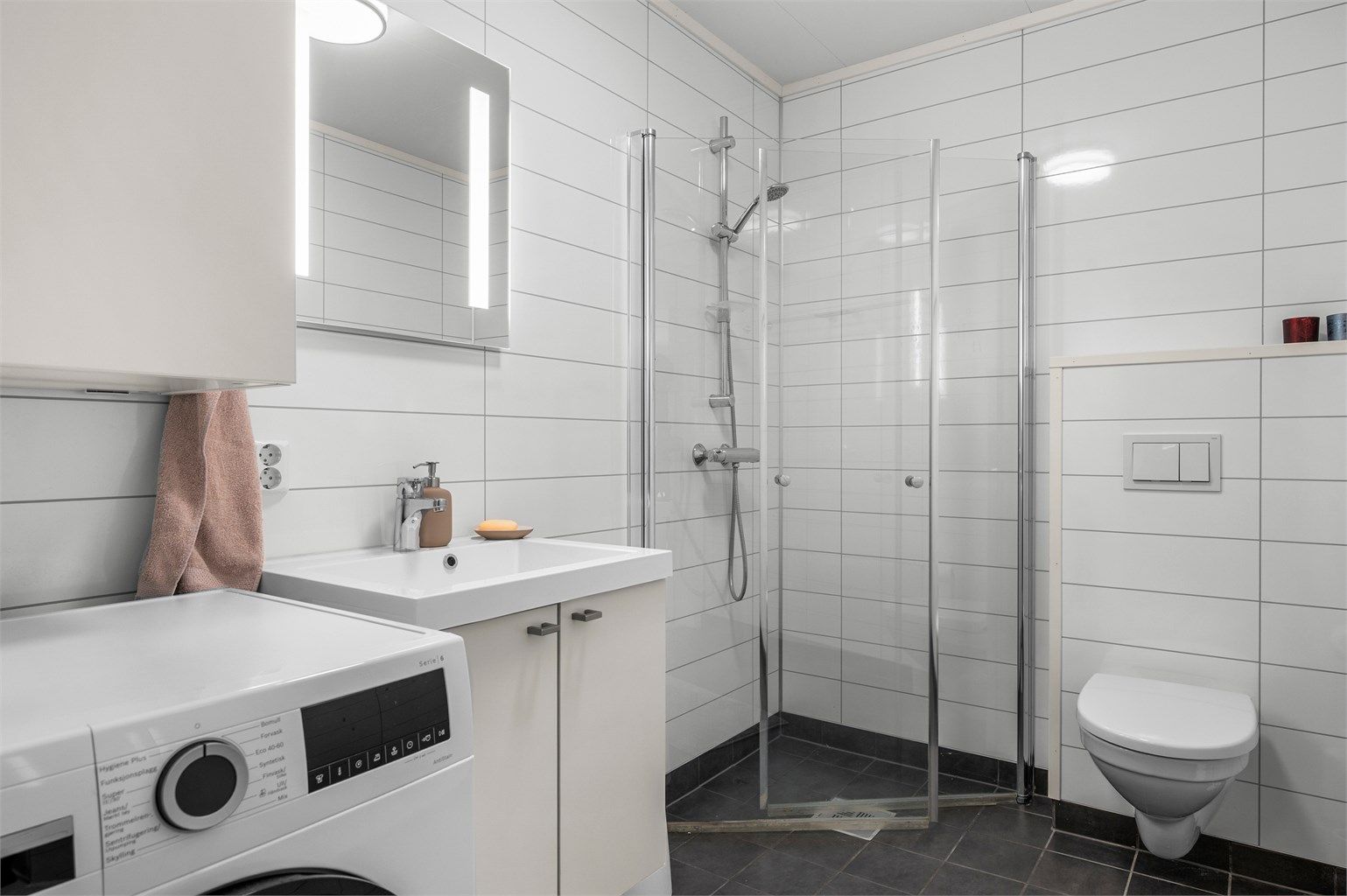 Badet i underetasjen er fra byggeår og har servantinnredning, dusjhjørne, vegghengt toalett og opplegg for vaskemaskin.