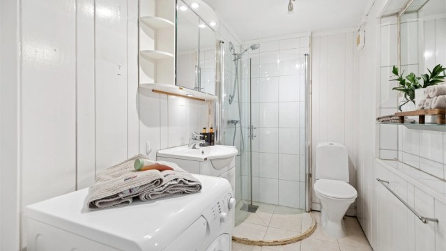 Hybel: Badet har fliser på gulv med gulvvarme. Dusjhjørne med innfellbare dører, lys servantinnredning, gulvstående toalett og opplegg for vaskemaskin.