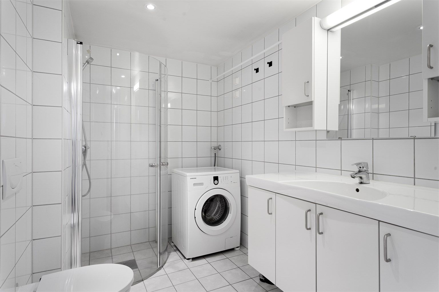 Badet har vegghengt toalett, dusjhjørne og vaskemaskin.