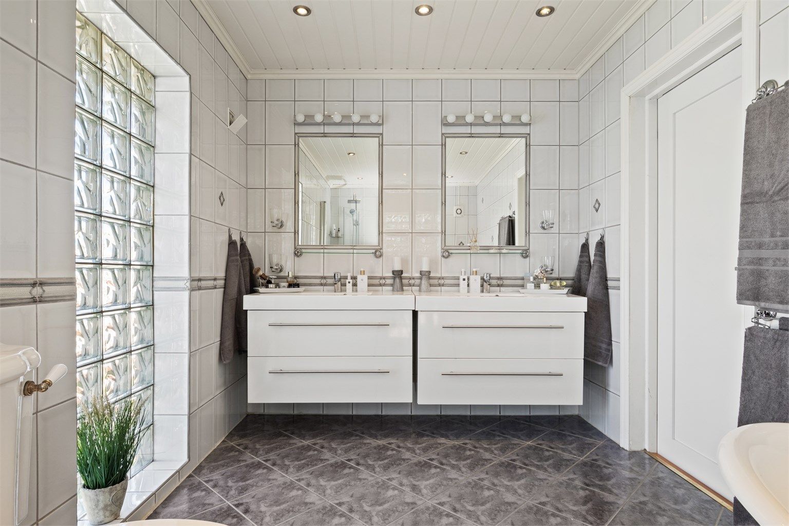 Videre er det et toalett i retrodesign, to servanter på hver sin hvite skuffeseksjon og to fastflisede speil med overlys.